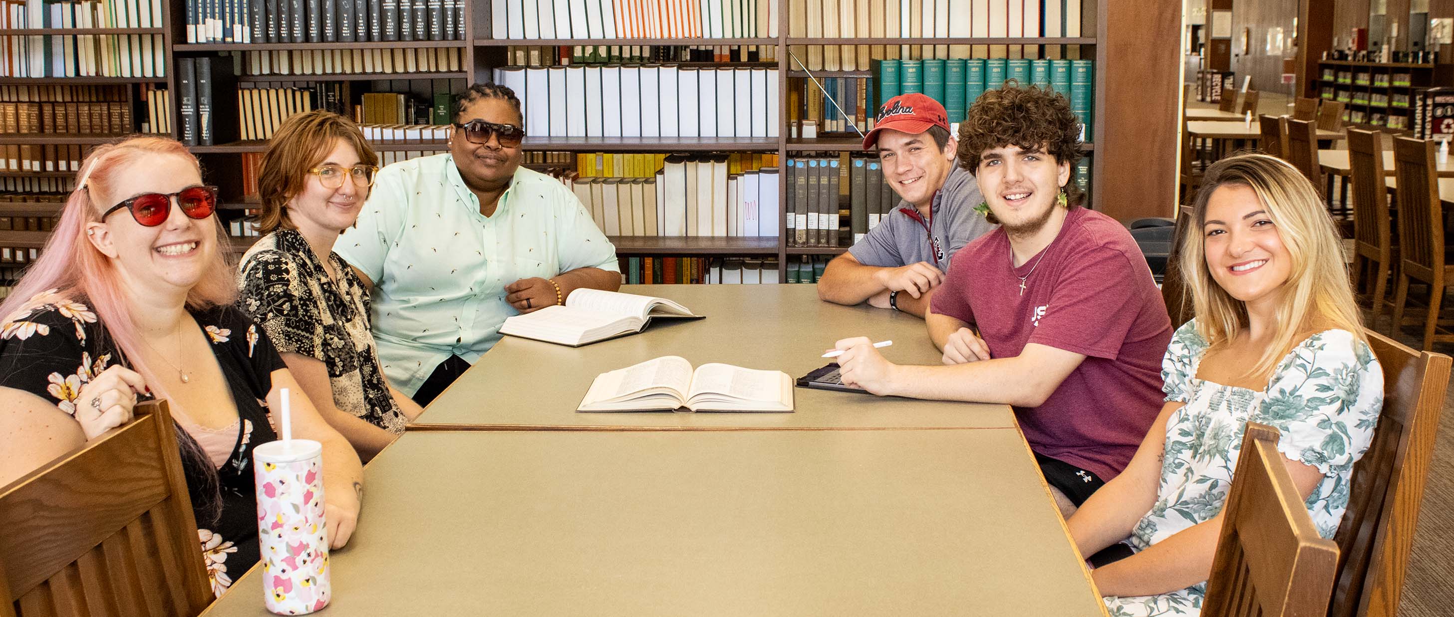 A group of students sit at a long wooden table in the library, with two books propped open and a stylus in one's hand.