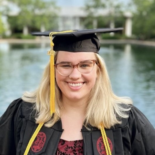 Young woman with blonde hair and glasses posing in black graduation cap and gown