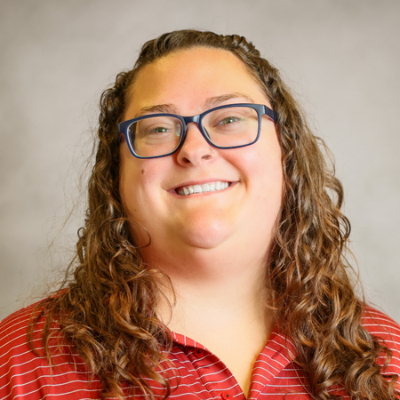 Professional headshot of a smiling person with long, curly brown hair wearing blue rectangular eyeglasses and a red striped collared polo shirt. The person is facing the camera against a soft gray studio background with warm, even lighting, creating a friendly, approachable, and professional appearance.