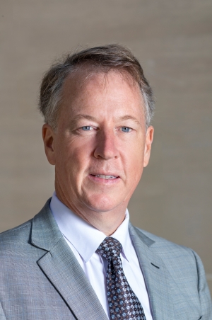 Professional headshot of Gerald A. McDermott wearing a light gray suit jacket, white dress shirt, and patterned tie, photographed against a neutral beige background.