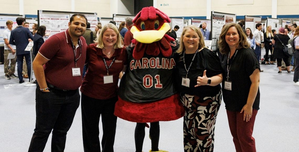 A group of four people, office of undergrdautea research staff, posing together indoors at Discover USC 2026, standing around Cocky, the University of South Carolina Gamecock mascot wearing a “Carolina 01” jersey. The individuals are smiling and wearing name badges, with rows of research posters and attendees visible in the background.