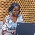 A smiling, seated person working on a laptop. The slightly angled, head-on view shows the top of the laptop, but not the screen.