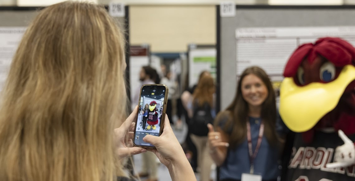 A Discover USC 2025 presenter snaps a photo of her co-presenter, who is posing with Cocky, both giving the spurs up hand signal.