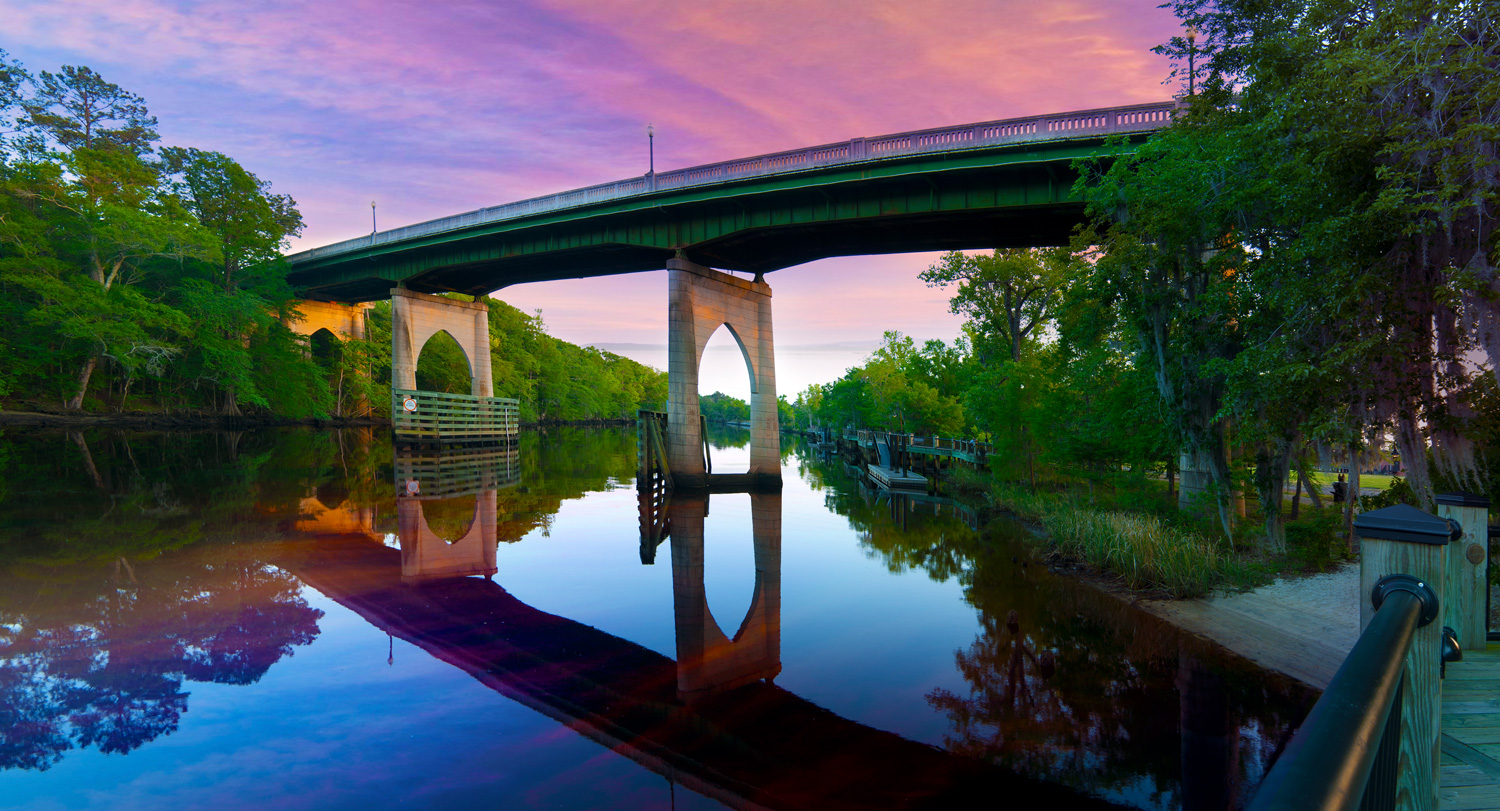 A green steel bridge with pointed stone arches spans a calm, tree-lined river beneath a pink and purple sunset sky, its supports reflected in the water.