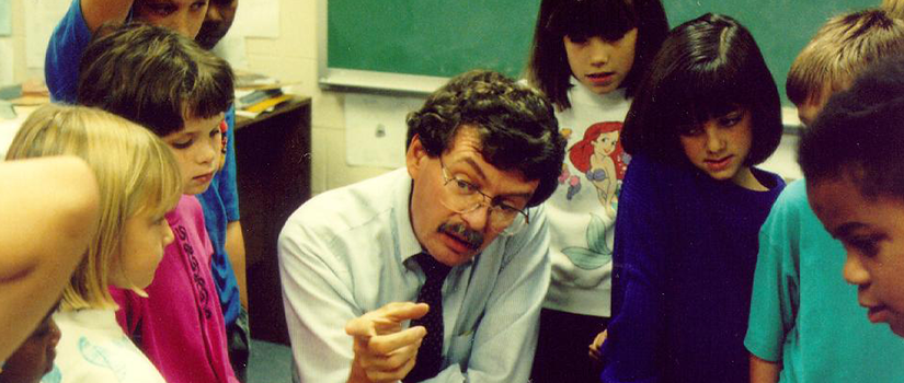 A teacher kneels in a classroom and shows a small object to a group of elementary school students gathered around him as they watch closely and listen.