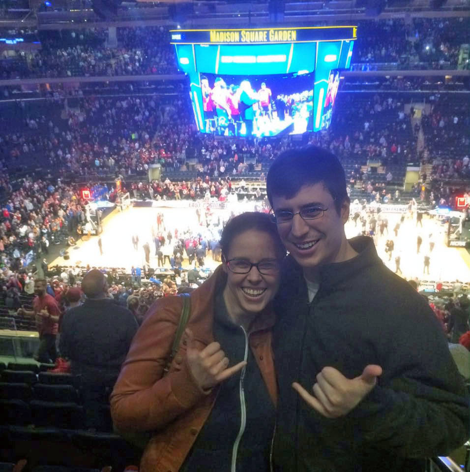 Two smiling fans pose with shaka hand gestures in the stands at Madison Square Garden during a basketball game. The court and a large digital scoreboard displaying the game are visible in the background.