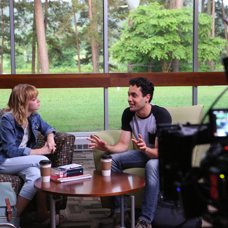 Actors Leah Merritt, “Sadie,” and Ian Gregg, “Ivy,” shoot a scene for Holy Irresistible. The actors are seated in what appears to be a library and are leaning toward each other, engaged in discussion