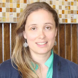 Annie wears a green oxford, navy sweater and silver earrings with her hair down in front of a wood and tile background