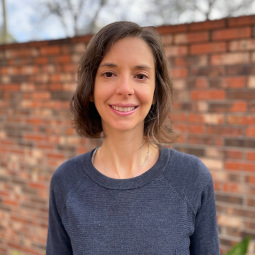 Carolyn wears a navy sweater, and is standing in front of a brick garden wall smiling