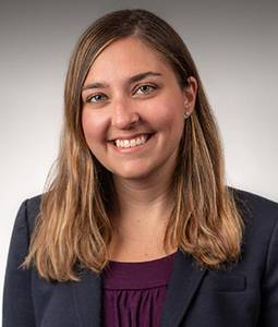 Emily Neger smiles at the camera, wearing a dark gray blazer over a purple blouse against a light gray background.