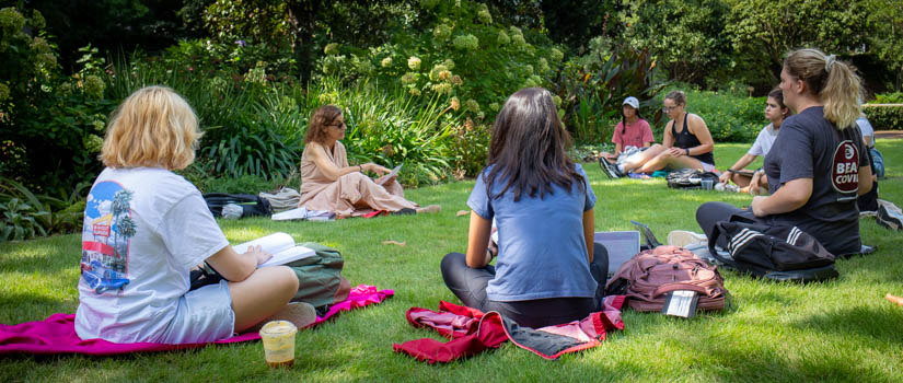 Three students holding notebooks standing in front of brick building. Tree in foreground.