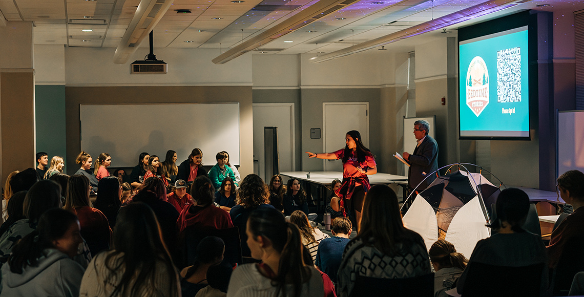 A woman telling a story by standing in the middle of a group of students sitting on the floor.