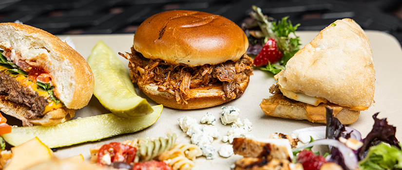 An assortment of food including a cheeseburger, pulled pork sandwich, pickles, and grilled chicken.