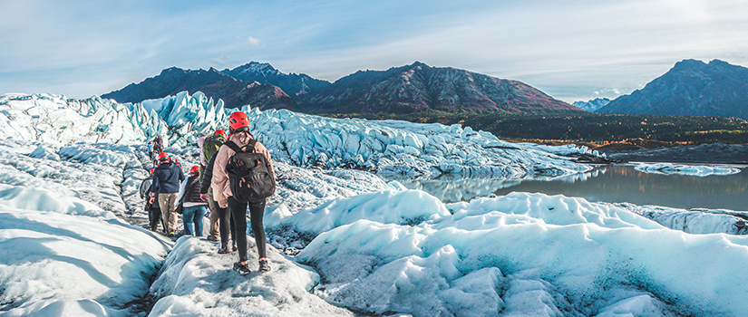 Group of hikers in helmets and winter gear trekking across a glacier with rugged mountains and blue sky in the background.