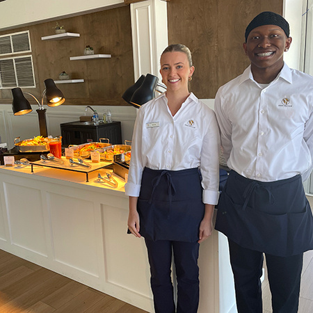 Two students working at a golf tournament pose for a photo by a buffet.