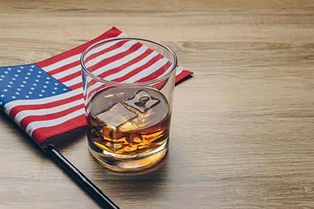 Glass of whiskey with ice resting on a wooden surface beside a folded American flag.