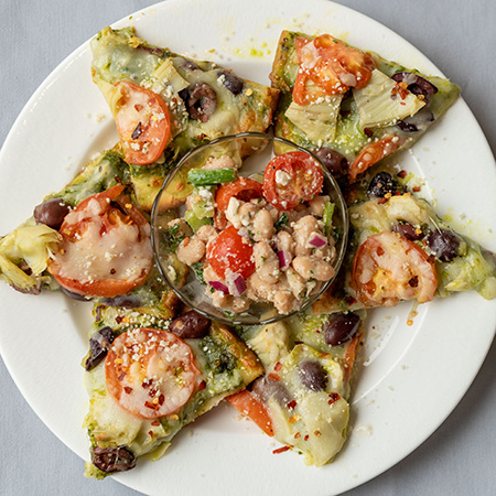 An overhead view of pita bread slices baked and topped with tomatoes, cheese and other vegetables along with a bowl in the center villed with tomatoes, pinto beans, diced onions and other veggies.