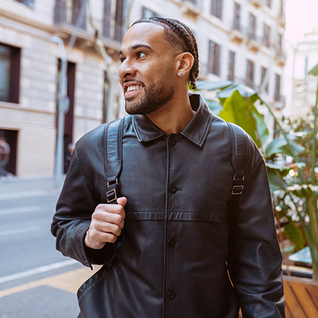 Smiling man wearing a black jacket and backpack walking through a city street lined with plants and buildings.