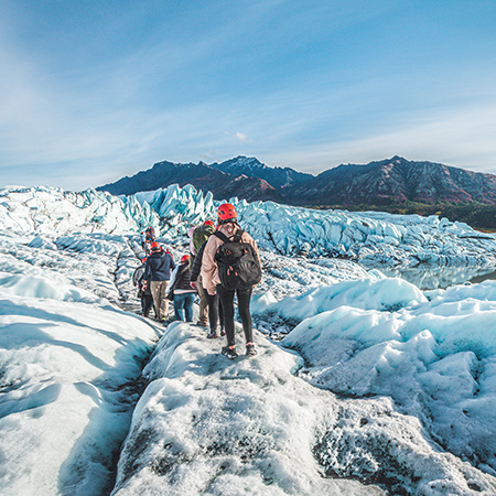 Group of hikers in helmets and winter gear trekking across a glacier with rugged mountains and blue sky in the background.