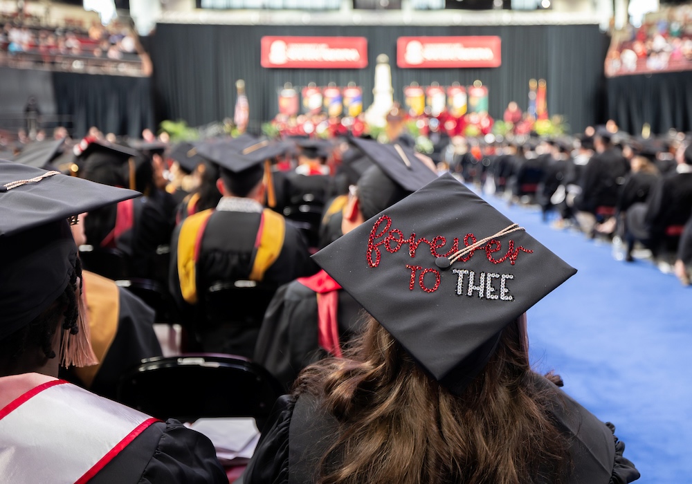 woman sitting in crowd wearing black graduation cap with glittery lettering that reads "forever to thee"