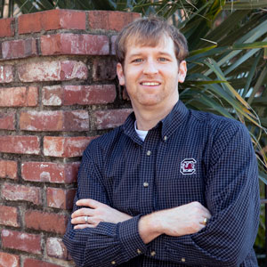 man leans against a brick wall with greenery in the background