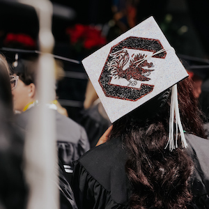 person in graduation robes wearing a bedazzled graduation cap with a gamecock and a block C on it 