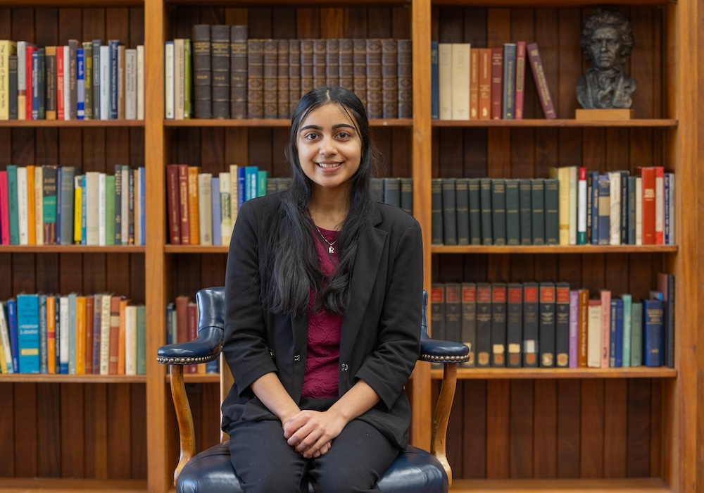 ria kothari in a black suit and garnet shirt sitting in front of a bookcase